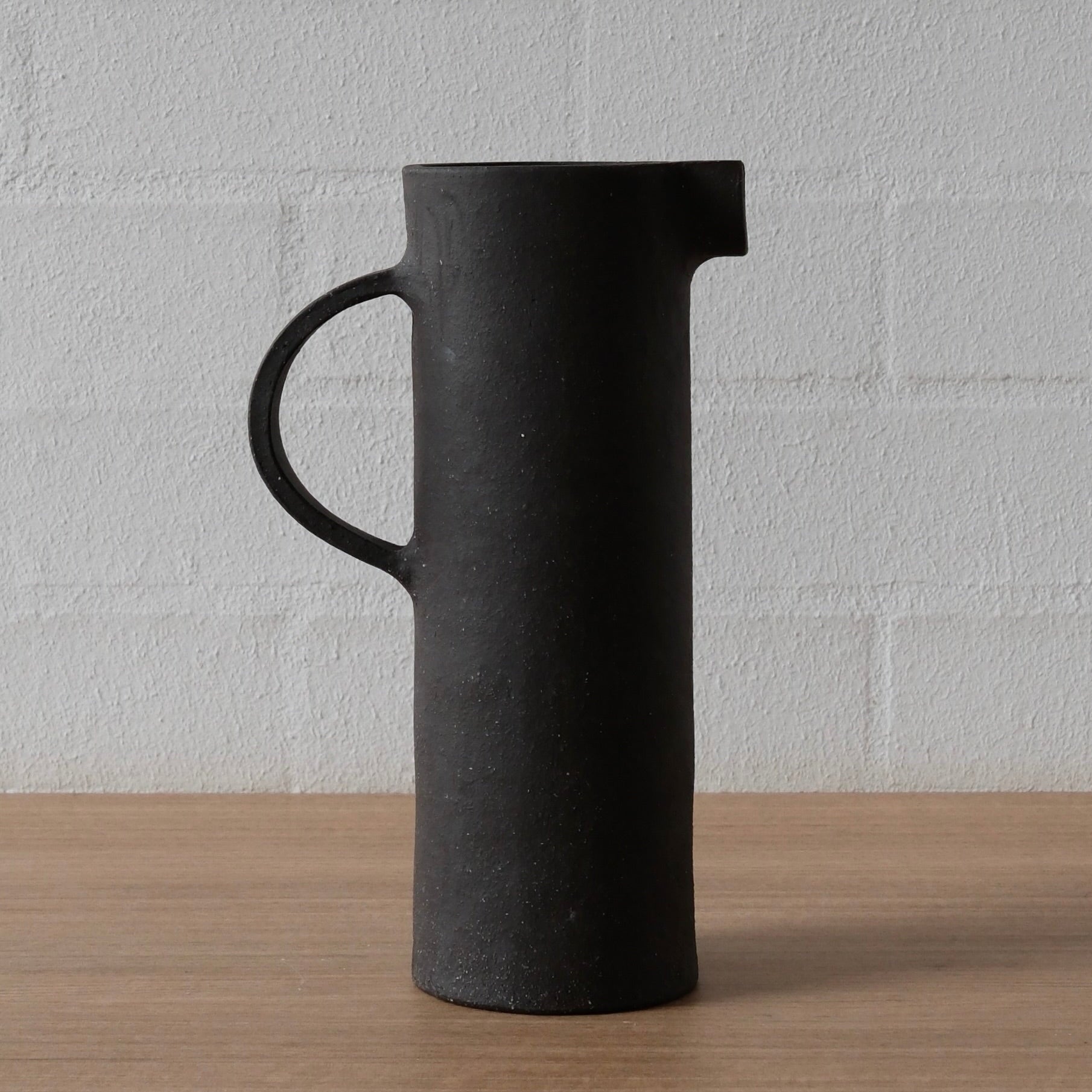 A matte black ceramic pitcher with a cylindrical body, a loop handle, and a small pouring spout, standing on a light wood surface against a white brick wall.
