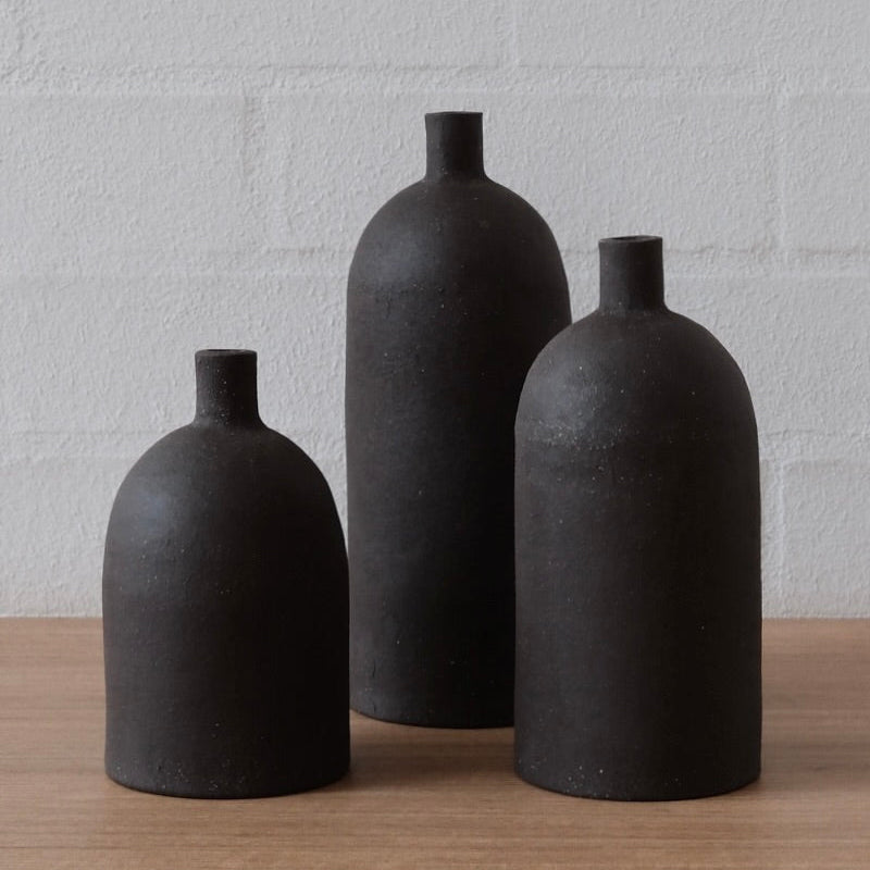 Three matte black ceramic bottle-shaped vases of different heights arranged on a light wood surface against a white brick wall.