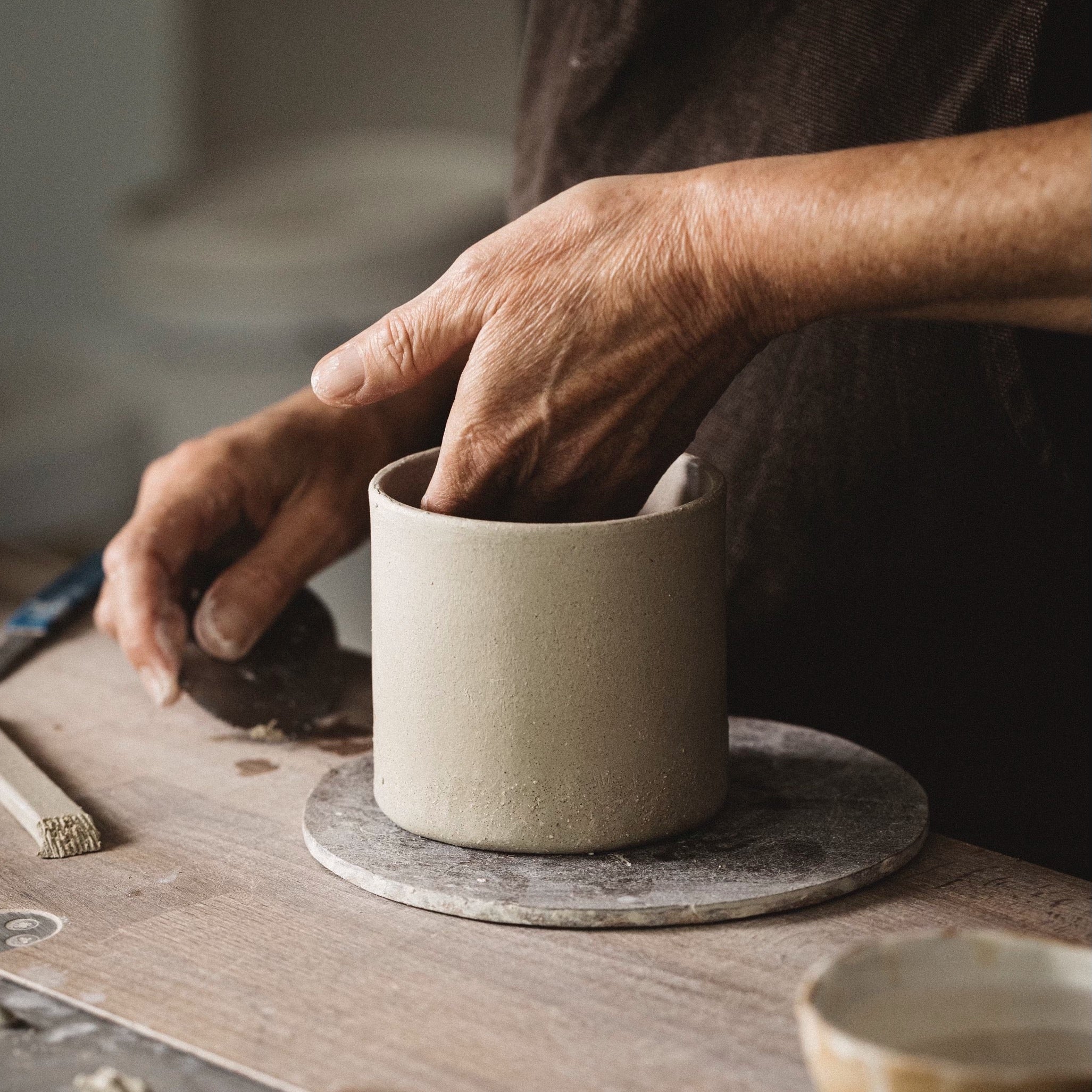 A person's hands shaping a ceramic cup on a round wooden board, with various crafting tools and materials scattered around the workspace.
